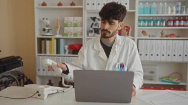 Klinikte çalışan genç bir adam, beyaz önlüklü bir adamla telefonda konuşurken profesyonel bir çalışma alanında dizüstü bilgisayar kullanıyor. Bu da sağlık hizmetlerine odaklanmış yoğun bir çalışma ortamı olduğunu gösteriyor..