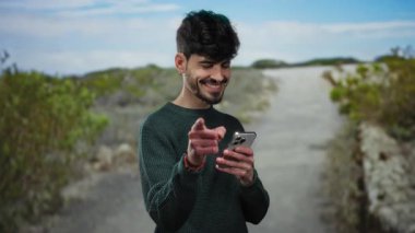 Genç İspanyol adam gülümseyip kameraya işaret ederken parkın dışında manzaralı bir arka planda telefon tutuyor, yeşil bir kazak giyiyor ve güneşli bir günün tadını çıkarıyor..
