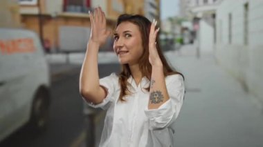 Kadın şehir caddesinde elleri ile çerçeve jesti, görünür dövmeli, genç ve rahat, açık havada gün ışığında günlük beyaz gömlek ile gülümsüyor.