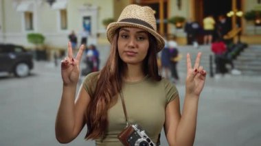 Kameralı ve şapkalı İspanyol kadın sokak ortamında barış işaretleri gösteriyor. Sıradan bir turist havası yayıyor..