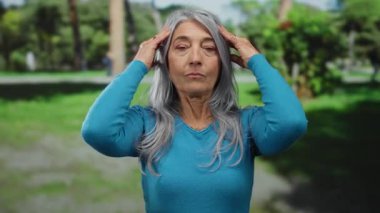 Gri saçlı, mavi tişörtlü son sınıf öğrencisi bir parkta duruyor, doğal ışık altında yemyeşil bir ortamda rahat ve sakin bir tavır sergiliyor..