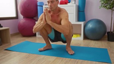 Genç adam spor salonundaki mavi paspasın üzerinde meditasyon yapıyor. Etrafı spor ekipmanlarıyla çevrili, huzurlu ifadeli ve odaklanmış pozlarla çevrili. Yumuşak ışıklandırma atmosferi altında.