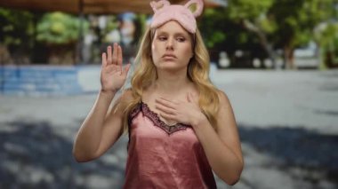 Pembe gece elbisesi ve uyku maskesi takmış genç bir kadın, elleri göğsünde, gün ışığında durgun bir poz veren yeşil ağaçlarla parkta duruyor..