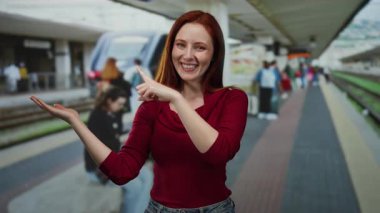 Tren istasyonunda gülümseyen, kırmızı giyen, elini gösteren, ayakta duran, dışarıda duran, kızıl saçlı, güzel, yetişkin, çekici, gösterişli seyahat ya da ulaşım sahnesi olan bir kadın..