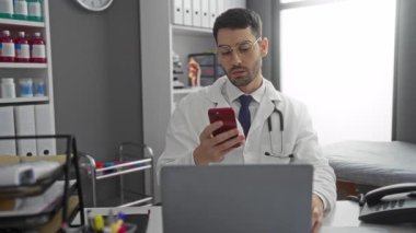 Klinikteki doktor cep telefonu kullandıktan sonra stresli görünüyor, arka planda tıbbi ekipmanlarla birlikte masadaki gözlükleri çıkartıyor ve erkek profesyonel sağlık hizmetlerine odaklandığını gösteriyor.