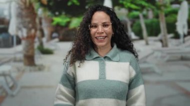 Güneşli bir sokakta gülümseyen İspanyol kadın, şakacı bir gülümsemeyle gözlerini kapatıyor ve gözlük takarak el kaldırıyor ve çizgili bir süveter giyiyor, açık havada neşe ve neşe yakalıyor..