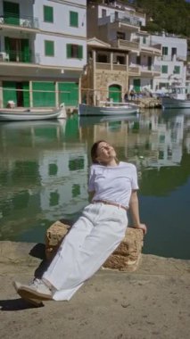 Cala figürlü Mallorca 'da güneşli Akdeniz limanında dinlenen bir kadın. Geleneksel tekne ve büyüleyici sahil mimarisi ile çevrili sakin deniz manzarasının tadını çıkarıyor..