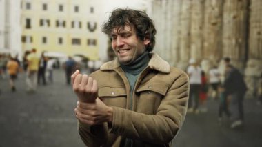 Yoğun şehir caddesinde kahverengi ceketli kıvırcık saçlı bir adam şehir ortamında bulanık yayalarla çevrili bileğini incelerken rahatsız görünüyor..