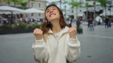 Şehir sokaklarında gülümseyen, beyaz bir süveter giyen, açık hava ortamında neşe ve mutluluk taşıyan, insanlar ve binalarla çevrili bir kadın..