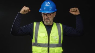 Siyah arka plana karşı kaslarını esneten, koruyucu iş ortamında inşaat özgüveni ve güvenlik farkındalığı gösteren sert şapka ve yansıtıcı yelek giyen bir adam..
