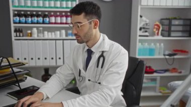 Klinikte kolları çaprazlanmış, gözlük takmış ve steteskop takmış genç İspanyol erkek doktor, iyi organize edilmiş tıbbi ofis ortamında masada oturuyor, profesyonelliği aktarıyor..