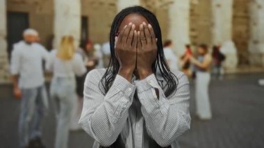 Açık şehir ortamında neşeli ifadeli gözlüklü bir kadın ve güneşli bir günde arka planda yürüyen insanlar..