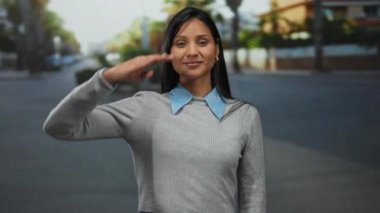 Mavi yakalı, gri kazaklı genç Latin kadın güneşli bir günde şehir ortamında askeri bir selam veriyor. Kendine güvenini ve disiplinini ifade ediyor..