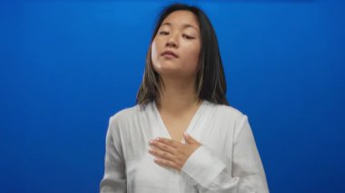 Genç bir kadın, mavi bir arka plana karşı yeminli bir jest yapıyor, beyaz bir gömlek giyiyor, bir stüdyo ortamında samimiyet, ciddiyet ve saygı tasvir ediyor..