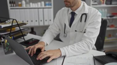 Klinikte beyaz önlük ve steteskop giyen genç bir adam, modern profesyonel ortamdaki tıbbi malzeme ve belgelerle çevrili bir masada otururken dizüstü bilgisayara odaklanmış.