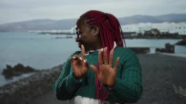 Kırmızı örgülü bir kadın arka planda okyanus kıyısında yeşil kazak giyip duygularını dışa vurarak, duygularını dışa vurarak reddediyor..