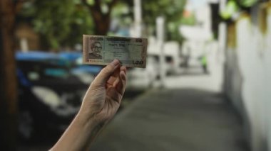 Bangladeş banknotunu elinde tutan adam, ağaçlar ve arabalarla dolu bir caddede canlı bir şehir manzarası yaratıyor.