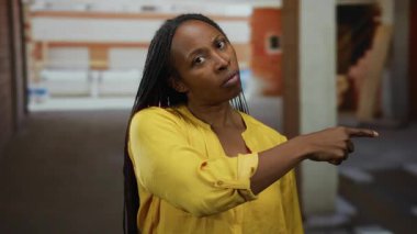 Bitmemiş bir endüstriyel binada, üzerinde ciddi bir ifade olan sarı bir tişört olan bir inşaat alanını işaret eden bir kadın.