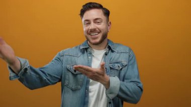 Kot ceketli sakallı genç adam gülümsüyor ve ellerini turuncu bir arka plana doğru açık bir şekilde işaret ediyor.