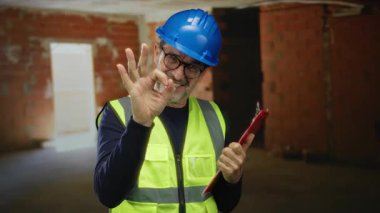 Güvenlik teçhizatı takmış yaşlı İspanyol bir adam, bina içi inşaat alanında elinde bir panoyla, kendine güvenini ve güvenini gösteriyor..