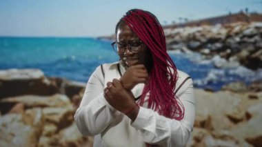 Pembe örgülü bir kadın, deniz kenarında acı içinde bileğini tutuyor turkuaz bir deniz ve kayalık plajın arka planına karşı, dışarıda, dinlendirici ortamların ortasında rahatsızlığını dile getiriyor..