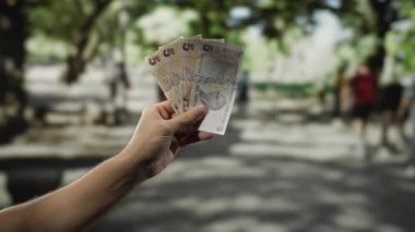 Bir şehir caddesinde elinde Türk lirası banknotları tutan adam bulanık insanlar ve ağaçlarla açık bir şehir ortamında mali işlem gösteriyor..