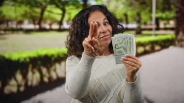 Bahçede Thai baht banknotları tutan bir kadın, 'seni izliyorum' jesti, şakacı bir güven ve doğal ortam ile dikkat çeker.