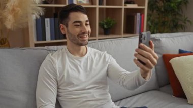 İspanyol bir adam rahat bir şekilde modern bir oturma odasında oturmuş, rahat ve mutlu bir ifadeyle, şık dekorlu rahat bir ev ortamını vurguluyor..