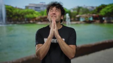 Siyah saçlı bir adam parkta gönüllü tişört giyiyor. Etrafı yemyeşil, umut dolu bir ifadeyle şükranlarını sunuyor ve ellerini sımsıkı tutuyor..