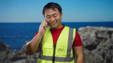 Güvenli bir yelek giyen bir adam deniz kenarındaki bir yerde akıllı bir telefonla konuşuyor. Okyanus ve kayaların arka planına gülümsüyor, iş ve açık hava aktivitelerini birleştiriyor..