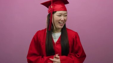 Kırmızı mezuniyet elbisesi giymiş, şapkalı, ağzı pembe arka planda gülen bir kadın başarıyı neşeyle kutluyor..