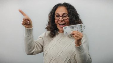 Orta yaşlı, gözlüklü bir kadın izole edilmiş beyaz arka plan üzerinde heyecanlı bir şekilde para taşıyor, sürpriz ve finansal başarıyı ifade ediyor..