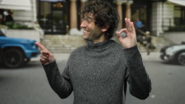Genç İspanyol bir adam gülümsüyor ve tek eliyle bir şehir caddesini işaret ederek neşeli ve olumlu bir doğa deneyimi gösteriyor..