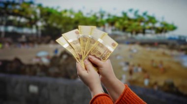 Macar paralarını sahil kenarında tutan kadın, görünür para birimleri ve deniz kenarı geçmişine sahip finansal veya seyahat kavramını vurguluyor..