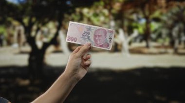 Güneşli bir parkta, arka planda ağaçlar olan bir yerde elinde Türk lirası banknot tutan adam doğal ortamda para gösteriyor..
