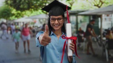 Diplomalı, gözlüklü ve mezuniyet şapkalı bir kadın şehir terasında başparmak kaldırıyor..