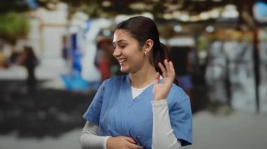 Açık havada gülümseyen İspanyol kadın hemşire, mavi önlük giyiyor, ağaçlarla kaplı bulanık bir şehir sokağına karşı kurulmuş, halka açık bir alanda sıcaklığı ve profesyonelliği aktarıyor..