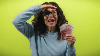 İzlanda Krona banknotları tutan kadın, neşeli bir şekilde uzaktaki sarı arka plana bakıyor, heyecan ve neşe dolu ifadeler sergiliyor..