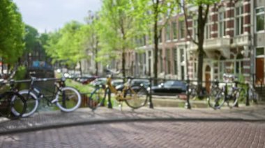 Amsterdam 'ın ikonik kanalının yanındaki kaldırımlı bir caddede bisiklet manzarası bulanık. Tarihi ağaçlar ve güneşli gökyüzünün altındaki büyüleyici tuğla binalar..