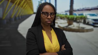 Şehir caddesinde kolları çapraz duran, gözlük ve sarı bluz takan, özgüven ve kararlılık saçan bir kadın..