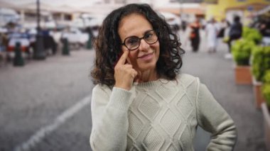 Açık hava ortamında, hareketli bir şehir caddesinde, gözlük ve süveter takmış, kıvırcık saçlı olgun bir güzeli gösteren bir gülümseme ile gözünü gösteren bir kadın..