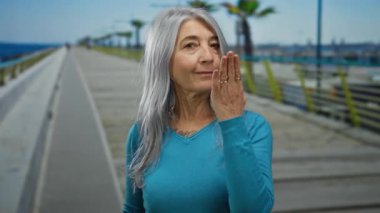 Gri saçlı yaşlı kadın deniz kenarında bir öpücük yolluyor, sakin bir kumsal sahili anını yakalıyor açık gökyüzünün altında rahat ve neşeli bir atmosferde..