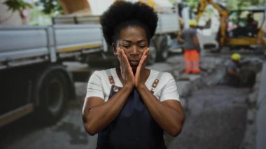 İnşaat alanında önlüklü bir kadın. Etrafı kamyon ve işçilerle çevrili. Düşünceli görünüyor..