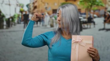 Gri saçlı kadın, canlı bir sokak ortamında yumruğunu kaldırmış pembe bir hediye kutusu tutuyor. Güç gösterisi yapıyor ve kutluyor..