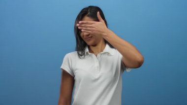 Beyaz tişörtlü genç Latin kadın, mavi arka plana karşı el ele hareketler yaparak yüzünü kapatıyor..