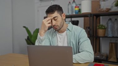 Ofisteki genç adam, masa başında dizüstü bilgisayarla otururken stresli görünüyor. Çalışma ortamını arka planda bitki ve raflarla gösteriyor..