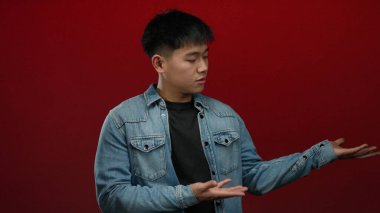 Kot ceketli genç adam, canlı kırmızı arka plana karşı ellerini yana doğru uzatıyor. Merakı uyandıran ya da duyguları sorgulayan..