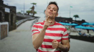 Kumsalda güneşli bir havada mavi şemsiyeler altında çizgili gömlek ve dövmeli yakışıklı bir adam.