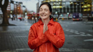 Parlak turuncu hapishane tulumlu kadın kalabalık şehir caddesinde duruyor, akşam üstü aydınlığında bulanık şehir geçmişiyle dışarıda duygularını ifade ediyor..