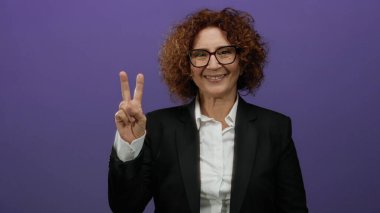 Gülümsemesi ve kendine güveni olan bir kadın canlı mor bir arka plana karşı barış işareti gösteriyor. Pozitif enerji ve iş kıyafetlerinde zarif bir tarz sergiliyor..